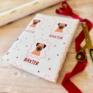 May include: White wrapping paper featuring a repeating pattern of a brown dog illustration inside a pink scalloped frame, accompanied by the red text BAXTER and small red stars. The paper is displayed with a roll, red ribbon, and gold scissors.