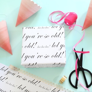 May include: White gift wrap with the text "lol you're so old!" repeated in black cursive writing. The text is surrounded by pink and white party hats and pink ribbon.