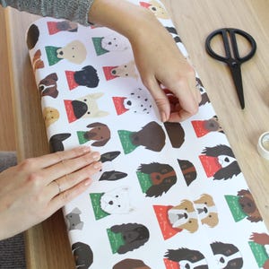 May include: White wrapping paper with a repeating pattern of colorful dog faces wearing Santa hats. A person is wrapping a gift with the paper.