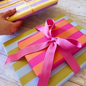 May include: Gift-wrapped presents with colorful striped paper. One package is wrapped in pink, orange, and white stripes and tied with a pink ribbon. Another is held in a hand and wrapped in yellow and purple stripes, tied with gold twine.