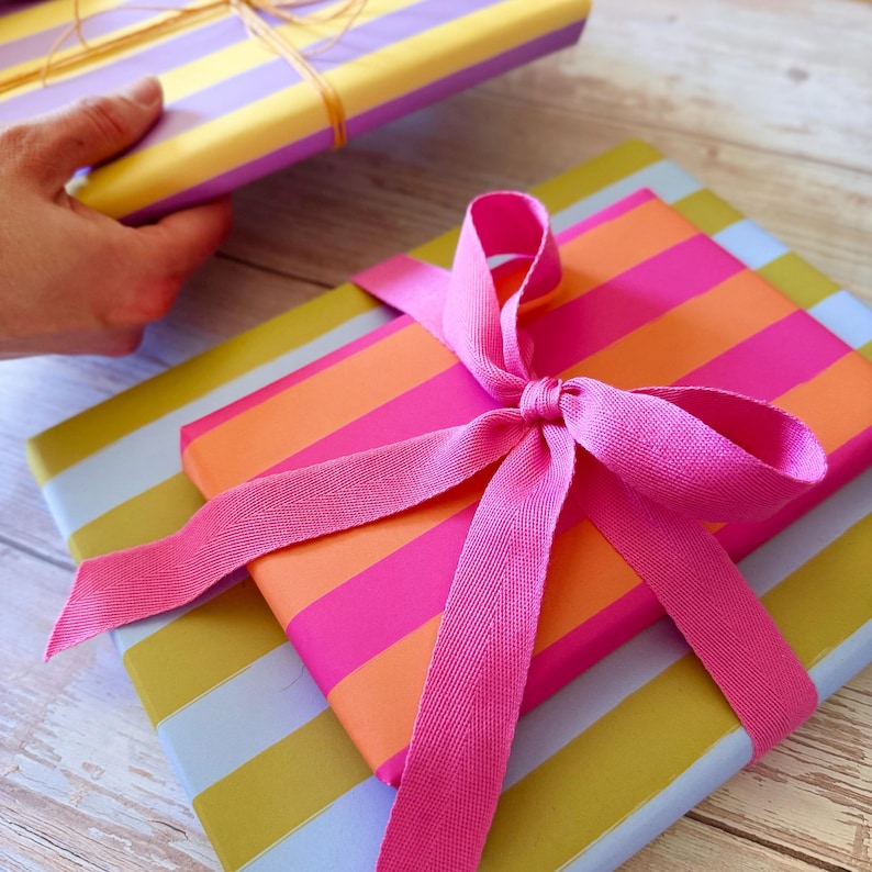 May include: Gift-wrapped presents with colorful striped paper. One package is wrapped in pink, orange, and white stripes and tied with a pink ribbon. Another is held in a hand and wrapped in yellow and purple stripes, tied with gold twine.