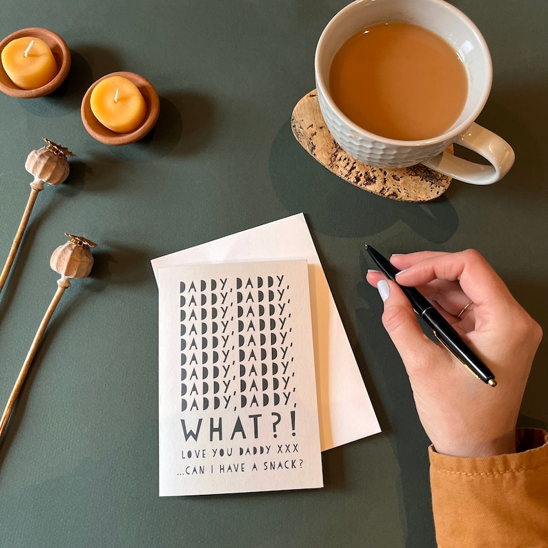 May include: A greeting card with the words "DADDY, DADDY, WHAT?!" in black text. The card is on a dark green surface, next to a white envelope, a cup of tea, and two small candles.