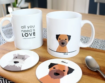 May include: Two white ceramic mugs with dog illustrations. One mug says "all you need is love...and a schnauzer". The other mug has a brown dog with the name "Meg" written below. Two round coasters with dog illustrations. One coaster has a gray dog and the other has a brown dog with the name "Meg" written above.