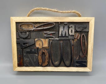 May include: A wooden frame with a string for hanging, containing a collage of vintage letterpress printing blocks. The blocks are arranged in a random pattern and include letters, numbers, and punctuation marks. The blocks are made of wood and metal and have a worn, aged appearance.