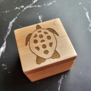 May include: A small, square wooden box with a turtle design etched on the lid. The turtle has a detailed shell pattern. The box is made of light-colored wood and has a simple, clean design.