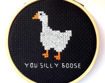Può includere: Un ricamo a punto croce in bianco e nero di un'oca bianca con zampe e becco arancioni. Il ricamo è incorniciato in un cerchio di legno e ha il testo "You silly goose" ricamato sotto l'oca.