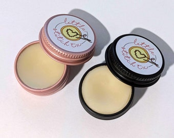 May include: Two small, round, metal tins with lids. The lids are labeled "little stitch fix" and feature a red heart with a needle and thread. The tins are open and contain a white, creamy substance.