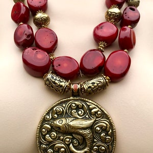 Puede incluir: Un collar de doble cadena con grandes cuentas de coral rojo y detalles dorados. El collar presenta un gran colgante redondo con un diseño intrincado de un pez en un patrón de remolino.