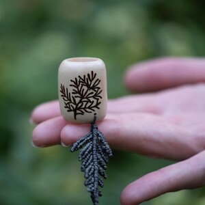 Puede incluir: Un pequeño tapón de botella de madera clara con un diseño de rama de árbol grabado en oscuro. Una hoja de macramé gris cuelga del tapón. El fondo es un verde suave y borroso.