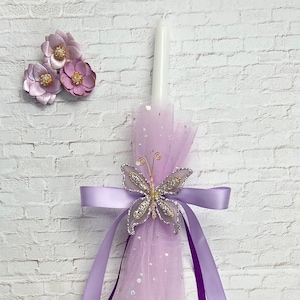 May include: A white candle decorated with a lavender tulle and a jeweled butterfly. Lavender ribbons and a cluster of lavender flowers complete the design. The candle is set against a white brick wall.