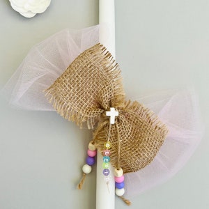 Könnte beinhalten: Eine weiße Kerze, verziert mit einer Schleife aus Jute, rosa Tüll und einem kleinen weißen Kreuz. Unter der Schleife hängt eine Kette aus pastellfarbenen Perlen. Links oben befindet sich eine weiße Stoffblume mit Strass-Mittelpunkt.