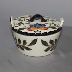 May include: A white ceramic lidded dish with a black handle and rim. The lid features a floral design with blue, yellow, and red flowers, and green leaves. The dish has two small handles and is decorated with green leaf patterns.