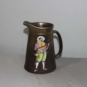 May include: A bronze-colored pitcher with a handle and spout. The front of the pitcher features a painted image of a musician in a white hat, purple jacket, and green pants, playing a lute. The pitcher has a dark brown background.
