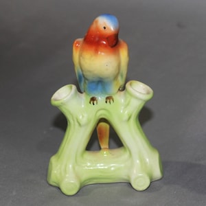 May include: A ceramic parrot figurine perched on a green tree branch with two small vases. The parrot is painted with red, yellow, blue, and green colors.