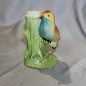 May include: A ceramic bud vase shaped like a tree stump, with a perched bird. The bird has a yellow head, blue chest, and brown wings. The vase is light green with a white interior.