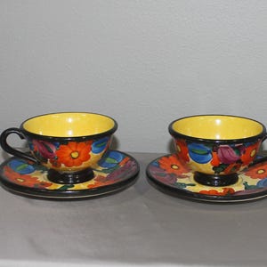 May include: Two ceramic teacups and matching saucers featuring a vibrant hand-painted floral pattern. The cups have bright yellow interiors, black handles, and bases, with exterior designs showcasing orange, blue, and purple flowers on a yellow background.