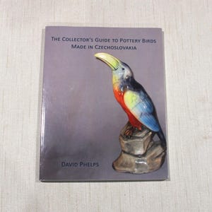 May include: A book titled "The Collector's Guide to Pottery Birds Made in Czechoslovakia" featuring a colorful ceramic toucan perched on a rock. The toucan has a yellow beak, red chest, and blue wings. The author's name, David Phelps, is printed at the bottom.