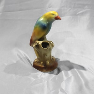 May include: A ceramic bird figurine perched on a tree stump. The bird has a yellow head, blue and green body, and a red beak. The tree stump is a light brown color. The figurine is likely a decorative item.