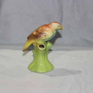 May include: A ceramic bird figurine perched on a green tree stump. The bird has a yellow beak, a brown and orange body, and a yellow tail. The tree stump is a bright green color. This decorative figurine is a charming addition to any home decor.