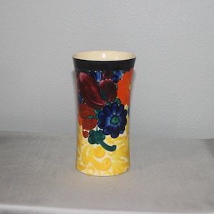 May include: A cylindrical ceramic vase with a cream-colored rim and a black band. The vase is decorated with a colorful floral pattern featuring red, blue, and orange flowers against a yellow background. The vase is approximately 8 inches tall.