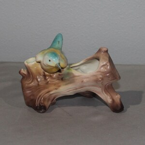 May include: A ceramic planter shaped like a brown tree log with a textured bark surface. A small bird with a blue head and yellow body is perched on the left side of the log. The interior of the planter is hollow and finished in a smooth white glaze.