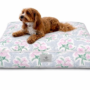 May include: A rectangular dog bed with a floral pattern in pink, green, and white on a light blue background. A brown dog with white markings is lying on the bed. The bed has a white border.