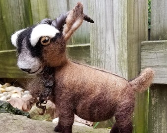 May include: A brown and black needle felted goat with white markings on its face. The goat is standing on a gray rock.