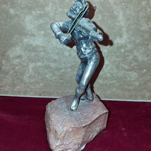 May include: A silver-toned metal figurine of a person playing a violin, depicted in a dynamic pose with one leg crossed over the other. The figure is mounted on a rough, rectangular, reddish-brown stone base with a textured, natural surface.