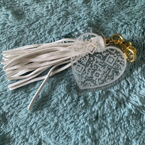 May include: A clear heart-shaped keychain with a white tassel. The heart has a QR code design and is attached to a gold metal ring.