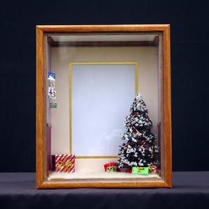Può includere: Una scena natalizia in miniatura in una cornice di legno. Un piccolo albero di Natale con neve bianca e bastoncini di zucchero rossi e bianchi si trova al centro della scena. Ci sono regali avvolti sotto l'albero e una cornice per foto sul muro dietro l'albero.