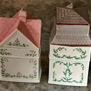 May include: Two ceramic house-shaped canisters with floral designs. The canister on the left has a pink roof and the canister on the right has a grey roof.