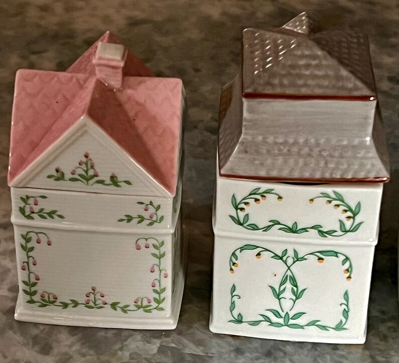 May include: Two ceramic house-shaped canisters with floral designs. The canister on the left has a pink roof and the canister on the right has a grey roof.