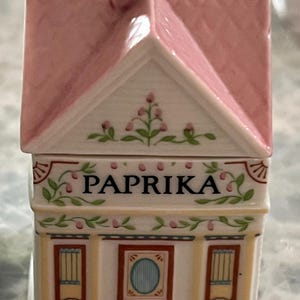 May include: A white ceramic house-shaped spice jar with a pink roof and floral details. The jar has a label that reads "PAPRIKA".