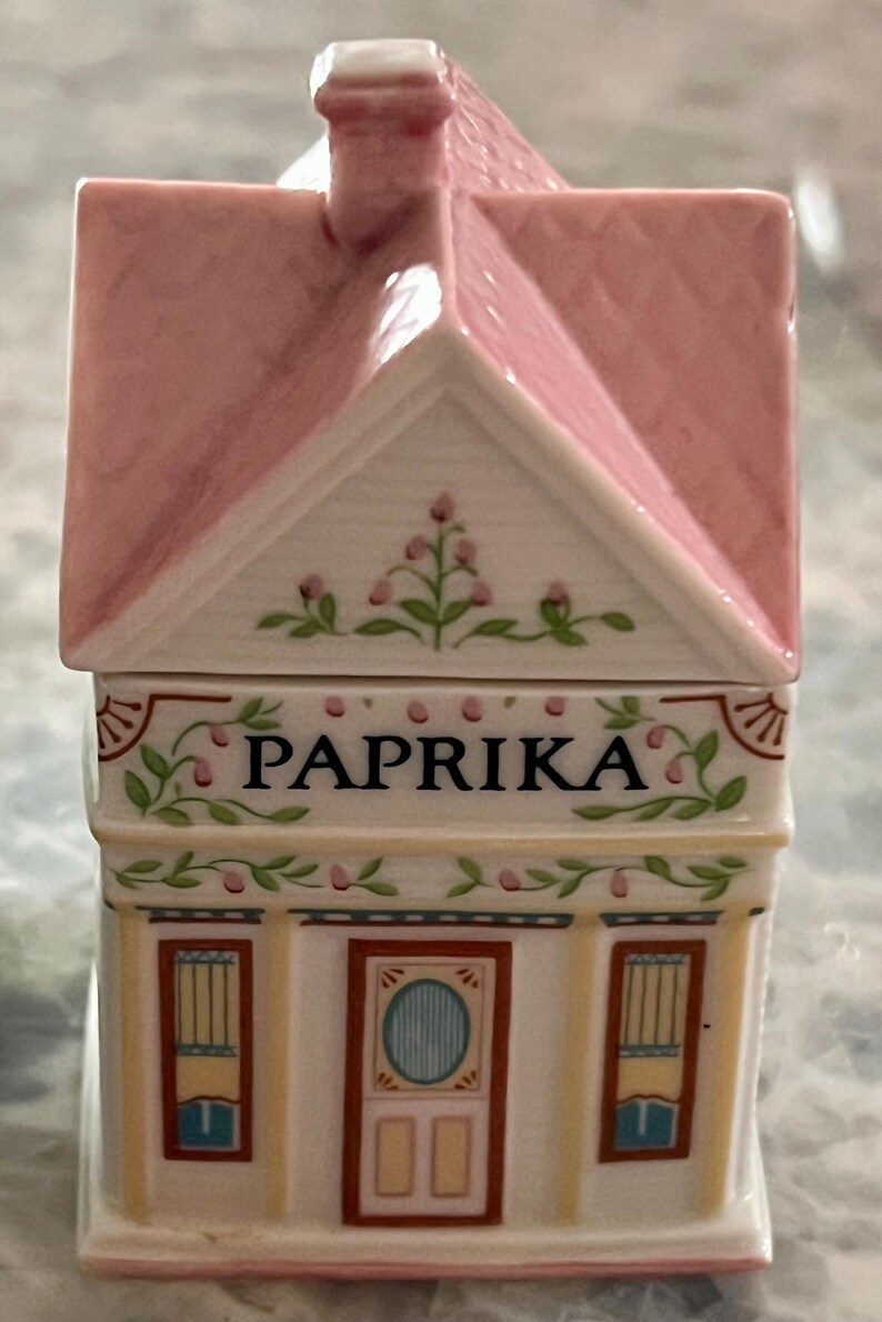 May include: A white ceramic house-shaped spice jar with a pink roof and floral details. The jar has a label that reads "PAPRIKA".