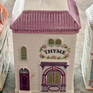 May include: A white ceramic spice jar shaped like a house with a pink roof and a sign that reads "THYME".