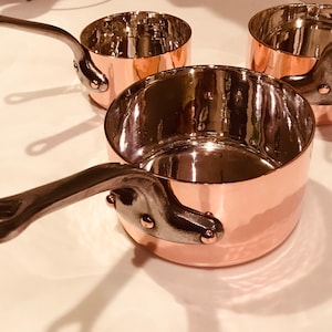 May include: Three copper saucepans with dark metal handles. The pans have a shiny interior and a hammered exterior. The handles are riveted to the copper body. The pans are sitting on a white surface.