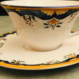 May include: A white teacup and saucer with a blue, yellow, and green floral design. The cup has a wide, rounded shape and a handle. The saucer has a scalloped edge.