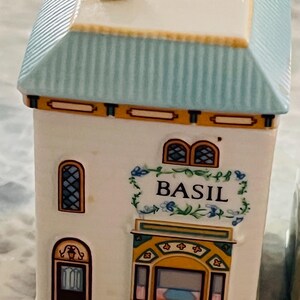 May include: A white ceramic spice jar shaped like a house with a blue roof and yellow trim. The jar has a window with a stained glass design and a sign that reads "BASIL".