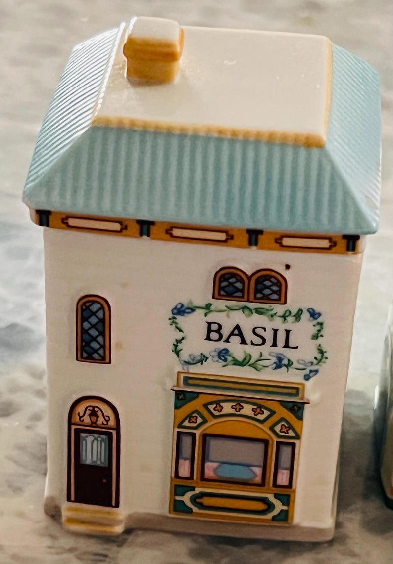 May include: A white ceramic spice jar shaped like a house with a blue roof and yellow trim. The jar has a window with a stained glass design and a sign that reads "BASIL".