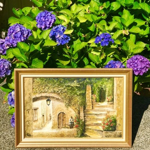 May include: A framed painting depicting a European street scene with a stone archway, steps, and a figure. The artwork features warm tones, a gold frame, and is set against a backdrop of vibrant purple hydrangeas.