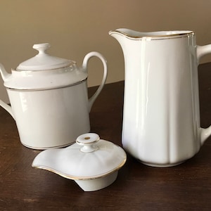 May include: A white porcelain sugar bowl with a lid and two handles, a white porcelain pitcher with a gold rim, and a white porcelain lid with a gold rim.