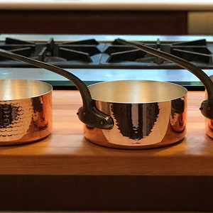 May include: Three copper saucepans with hammered surfaces and black handles. The saucepans are arranged in order of size, with the largest saucepan on the left and the smallest saucepan on the right.