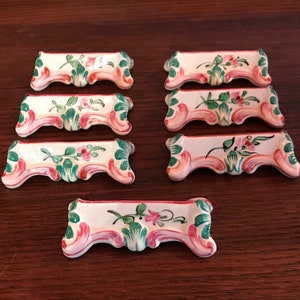May include: Set of seven white ceramic knife rests with pink and green floral designs. Each rest has a scalloped edge and a raised floral motif.