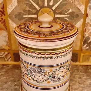May include: A white ceramic jar with a lid, decorated with hand-painted floral designs in shades of green, yellow, and brown. The jar has a traditional, rustic style.