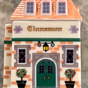 May include: A ceramic spice jar shaped like a house with a green door and two windows. The roof is orange with three windows. The front of the house has a sign that says "Cinnamon".