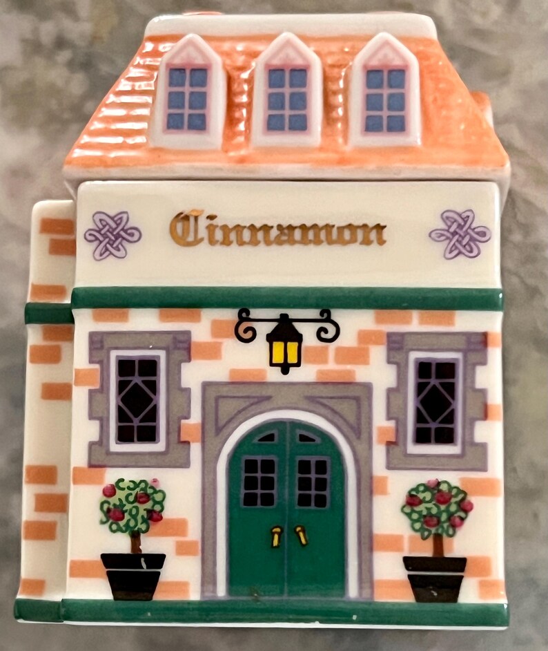 May include: A ceramic spice jar shaped like a house with a green door and two windows. The roof is orange with three windows. The front of the house has a sign that says "Cinnamon".