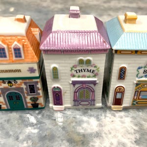 May include: Three ceramic spice jars shaped like houses with colourful roofs and painted details. The jars are labelled "Cinnamon", "Thyme", and "Basil".