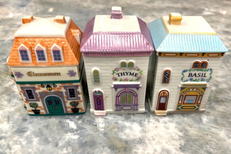 May include: Three ceramic spice jars shaped like houses with colourful roofs and painted details. The jars are labelled "Cinnamon", "Thyme", and "Basil".