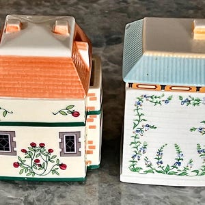 May include: Two ceramic canisters with floral designs. The canister on the left has a red roof and green trim. The canister on the right has a blue roof and yellow trim.