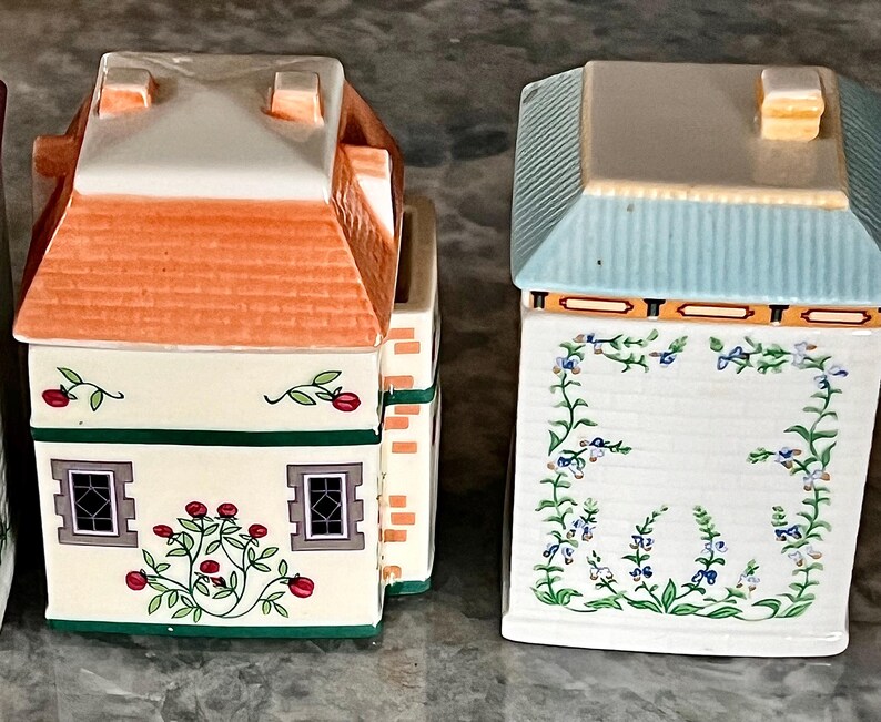 May include: Two ceramic canisters with floral designs. The canister on the left has a red roof and green trim. The canister on the right has a blue roof and yellow trim.